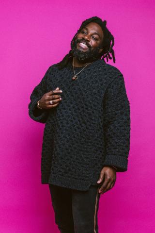 Author Jason Reynolds, wearing black knit sweater and pants, standing against a bright pink background. 