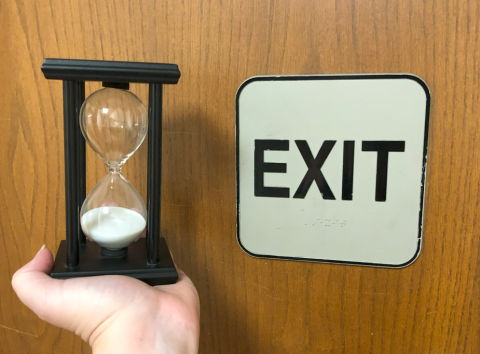 An hour glass is pictured next to a sign on a door reading "EXIT" in all caps and in braille.