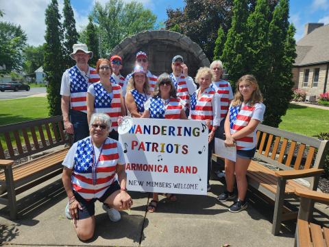 Group of harmonica players standing in a group wearing red, white, and blue vests behind a sign that reads "Wandering Patriots Harmonica Band New Members Welcome"