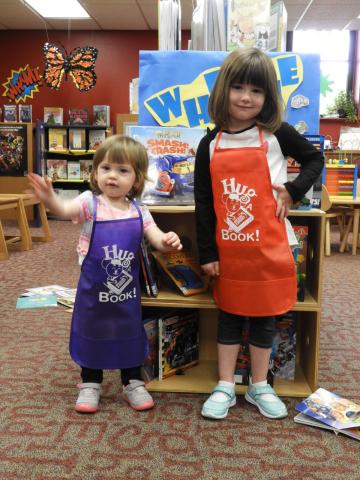 2 sisters wearing purple and orange children's art aprons with Hug-A-Book logo on front 