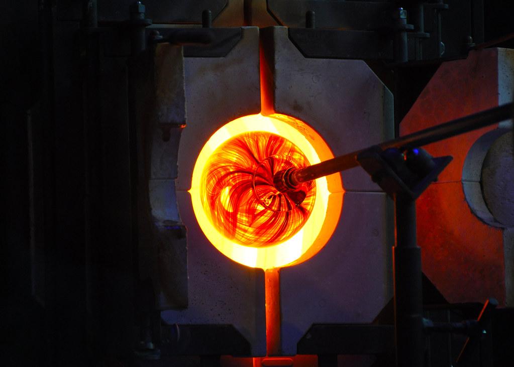 A glass ornament glows orange inside a furnace, and is held by a metal tool.