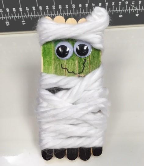 White yarn wrapped around popsicle sticks decorated with mummy face